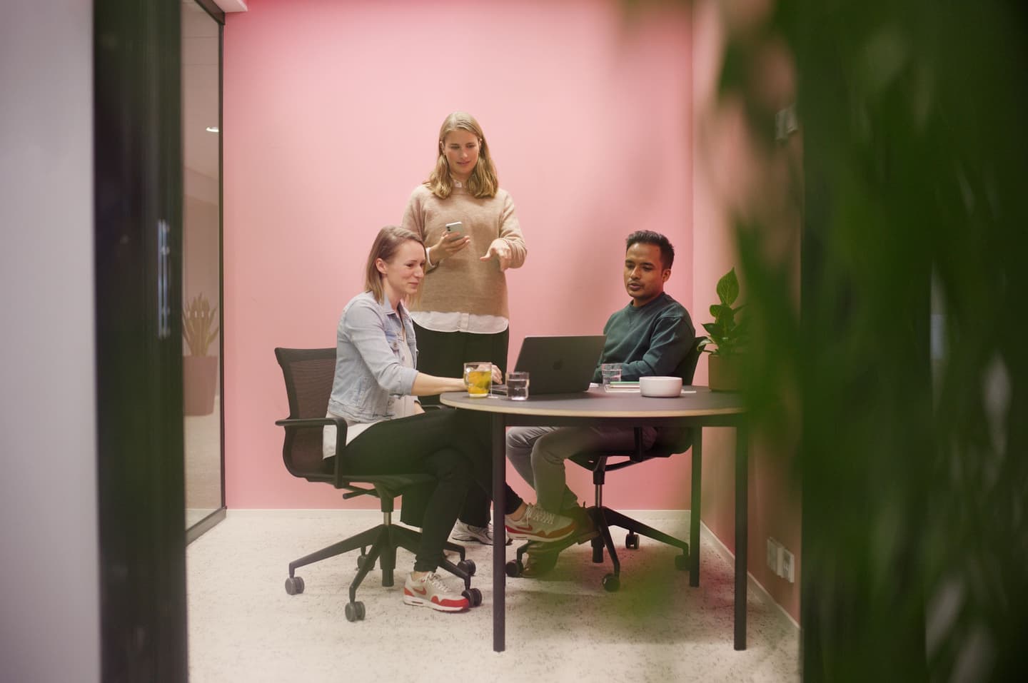 Three colleagues in a modern office meeting room with pink walls, one standing with phone while two sit at table with laptop.