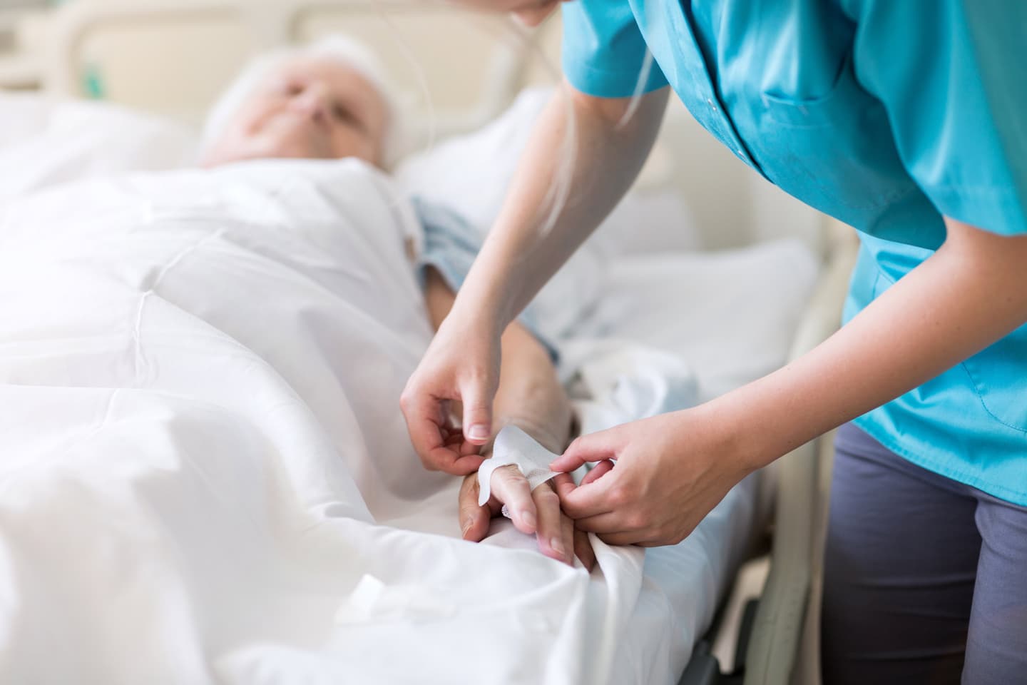 Healthcare worker in teal scrubs adjusting an elderly patient's IV or bandage in a hospital bed.