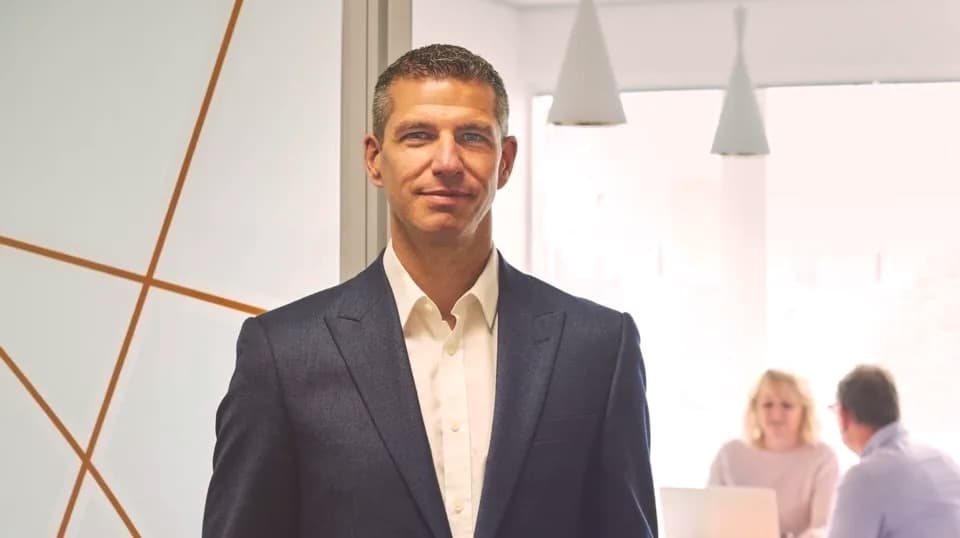 Professional in navy suit and white shirt standing in modern office with pendant lights and colleagues in background.