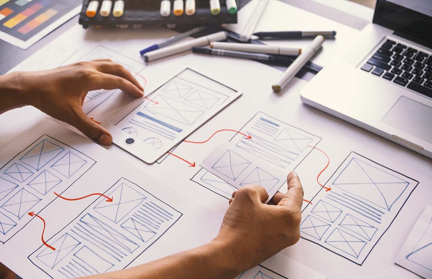 Hands working on website wireframe sketches with markers, laptop and design tools on a desk.