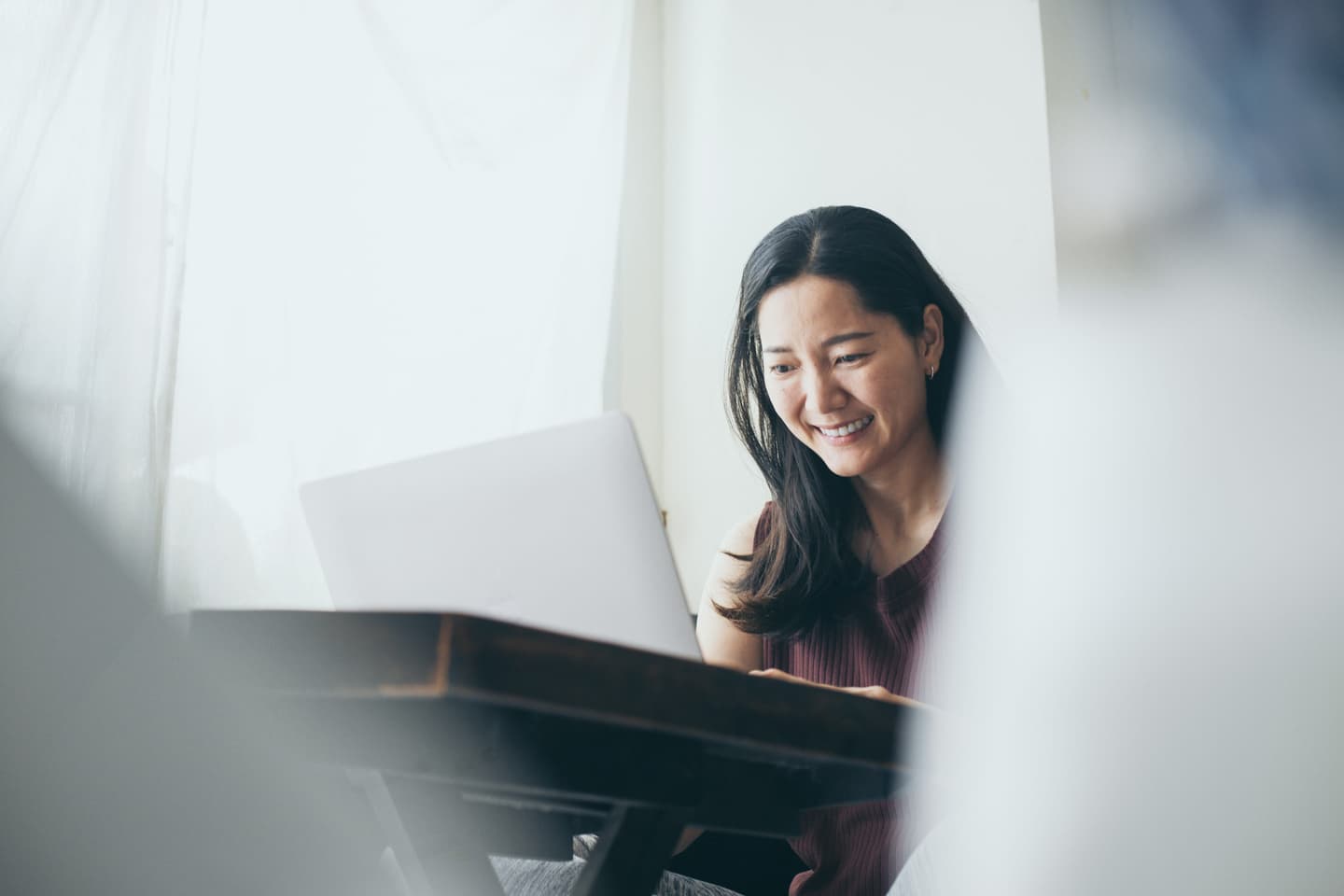 Person with long dark hair smiling while working on laptop near window in soft, natural lighting.