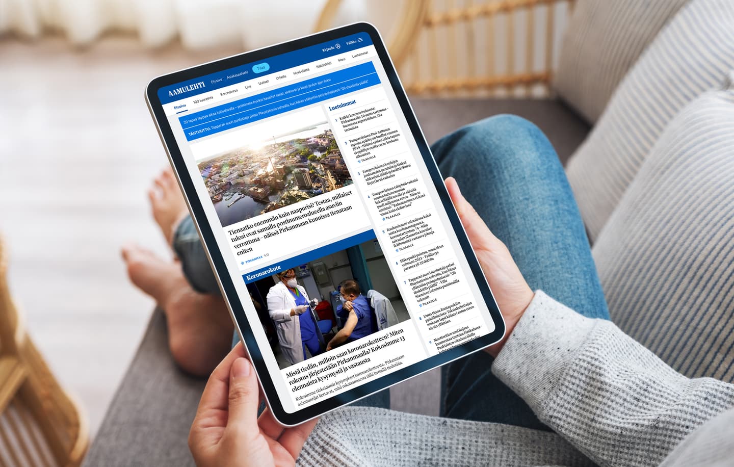 Person in grey sweater browsing Finnish news website on tablet, showing city view and healthcare articles.