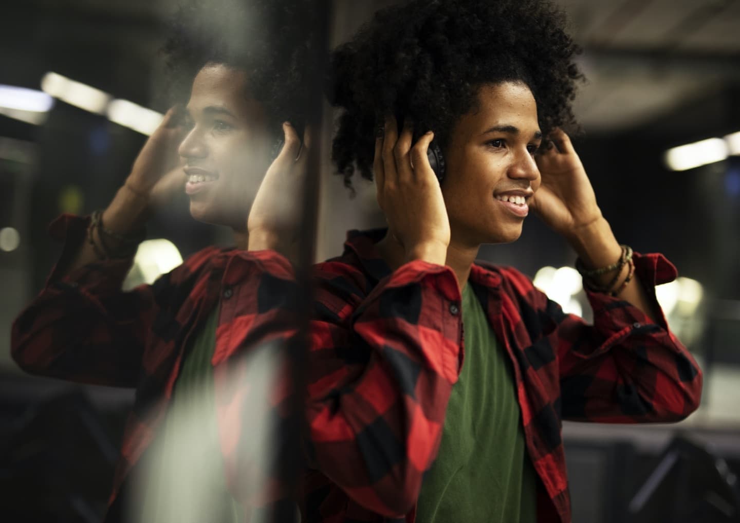 Person with curly hair wearing red plaid shirt and green top, smiling while adjusting headphones, with reflection visible.