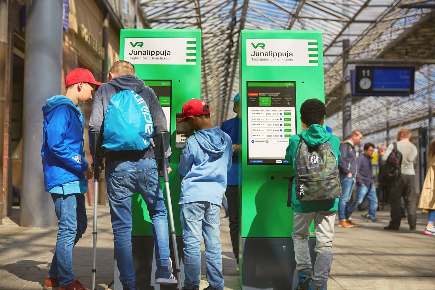 Young people using bright green VR train ticket machines at a station with glass roof overhead.
