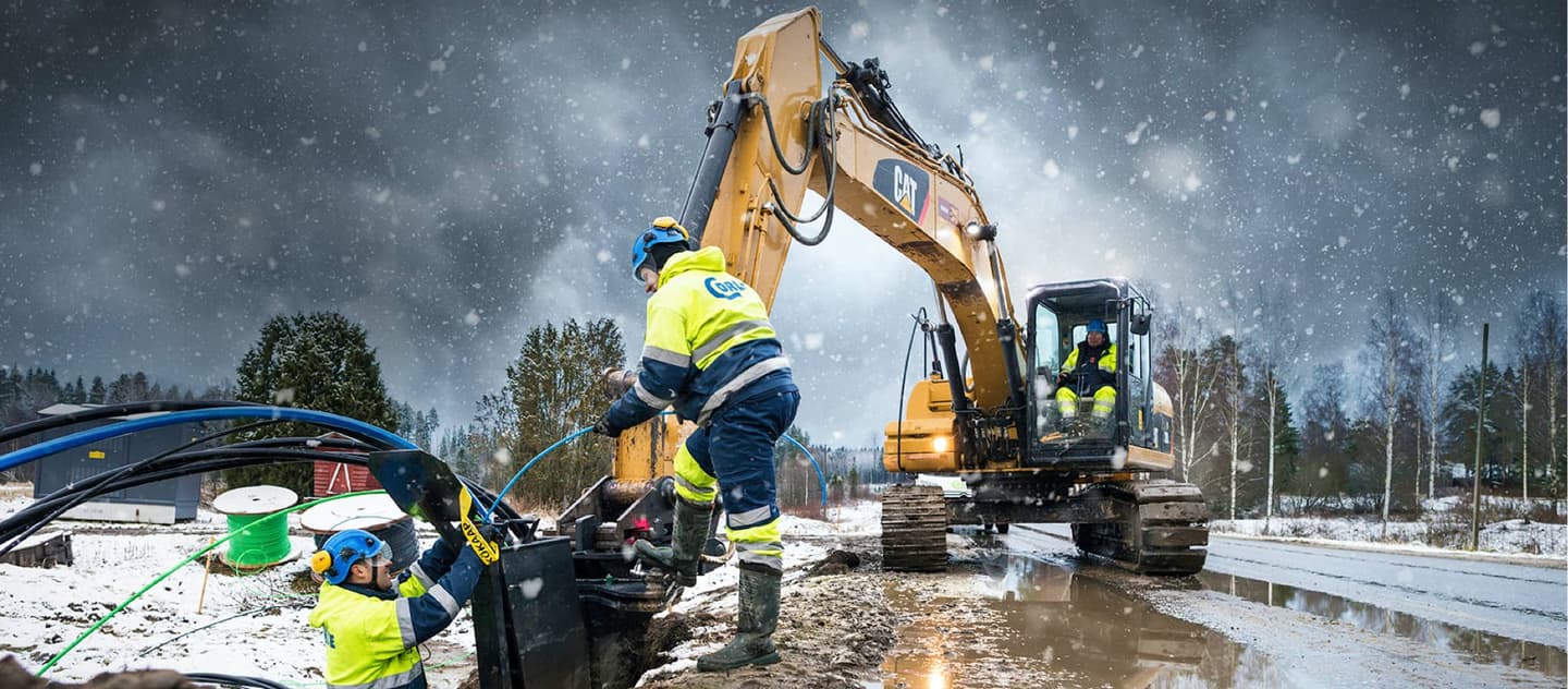 Construction workers in high-visibility gear operating a CAT excavator during snowfall at a muddy winter worksite.