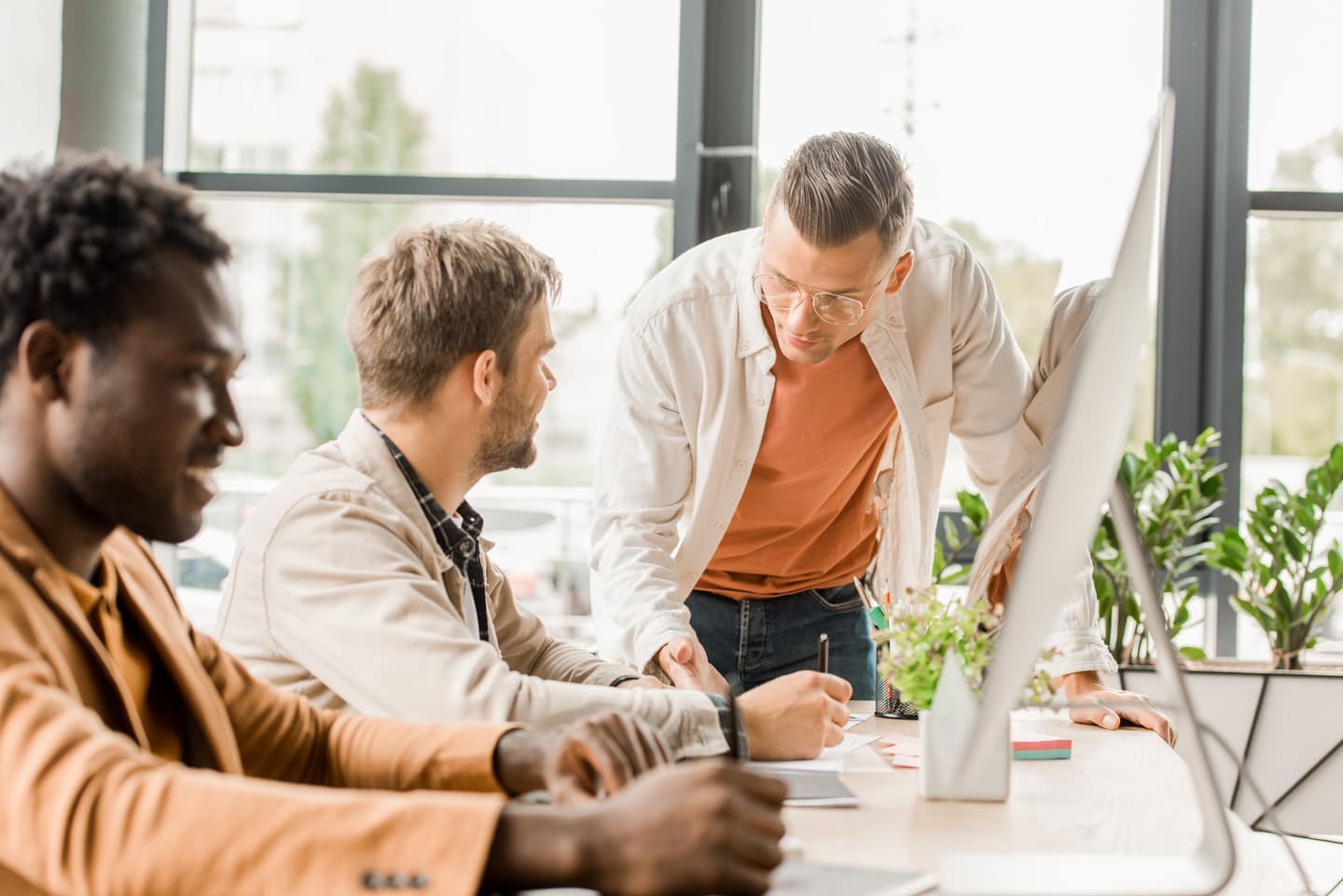 Three colleagues collaborating at a bright office desk with plants and a computer monitor, near large windows.
