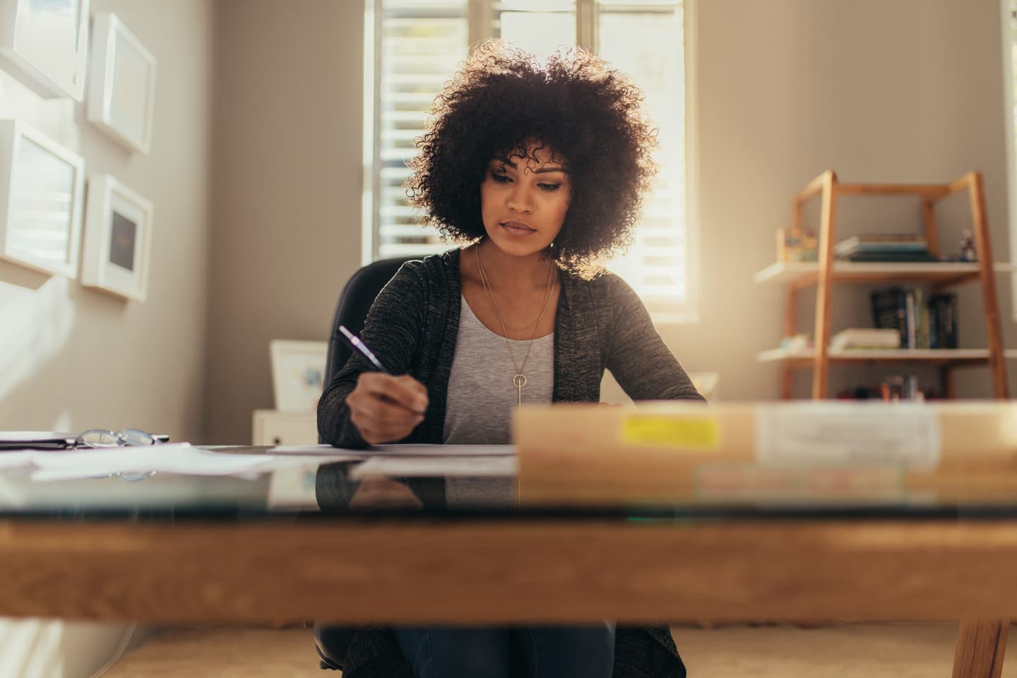 Person with curly hair writing at desk in bright home office with wooden bookshelf and window shutters in background.
