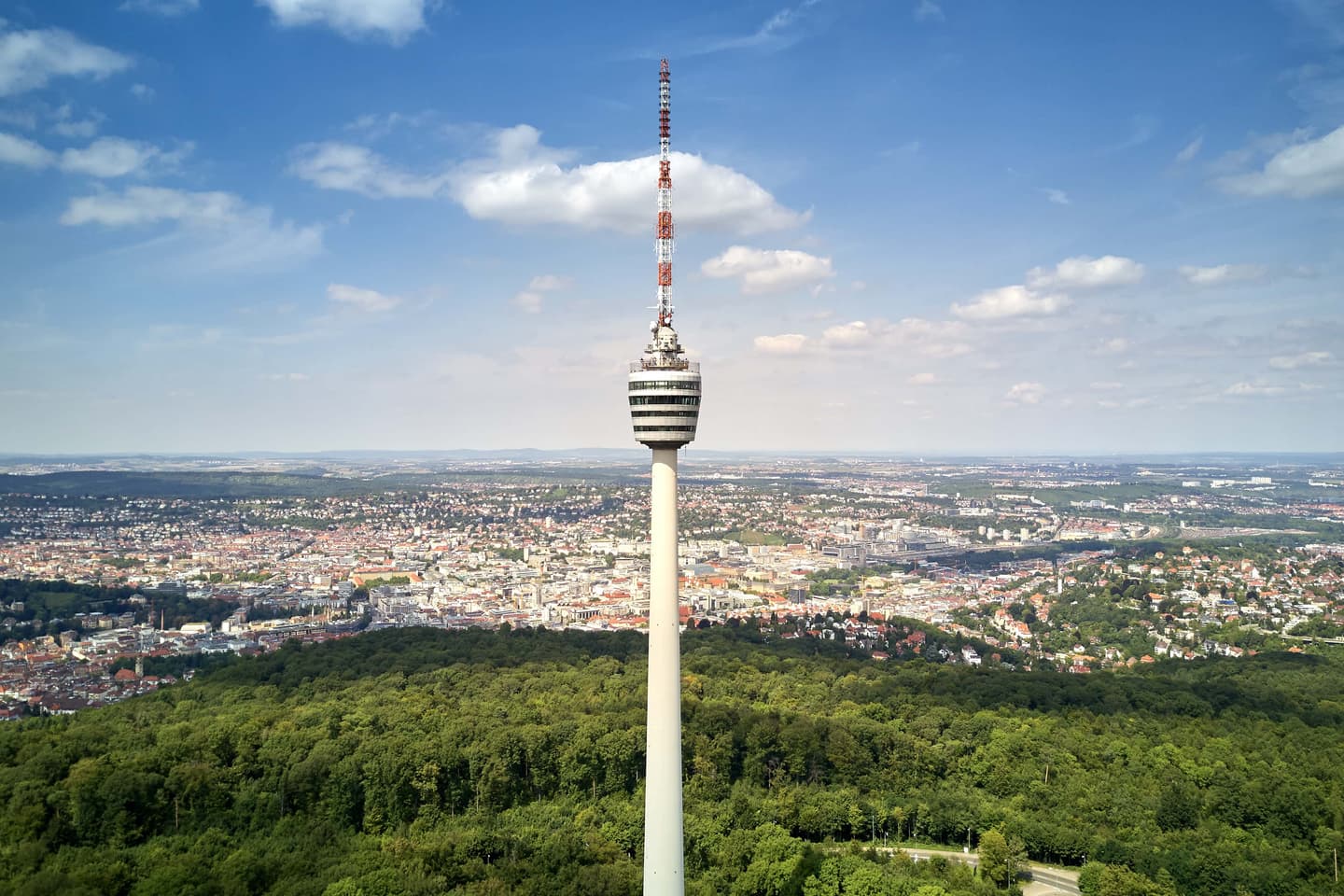 Tall television tower with striped observation deck rising above forest with cityscape view under blue sky with clouds.