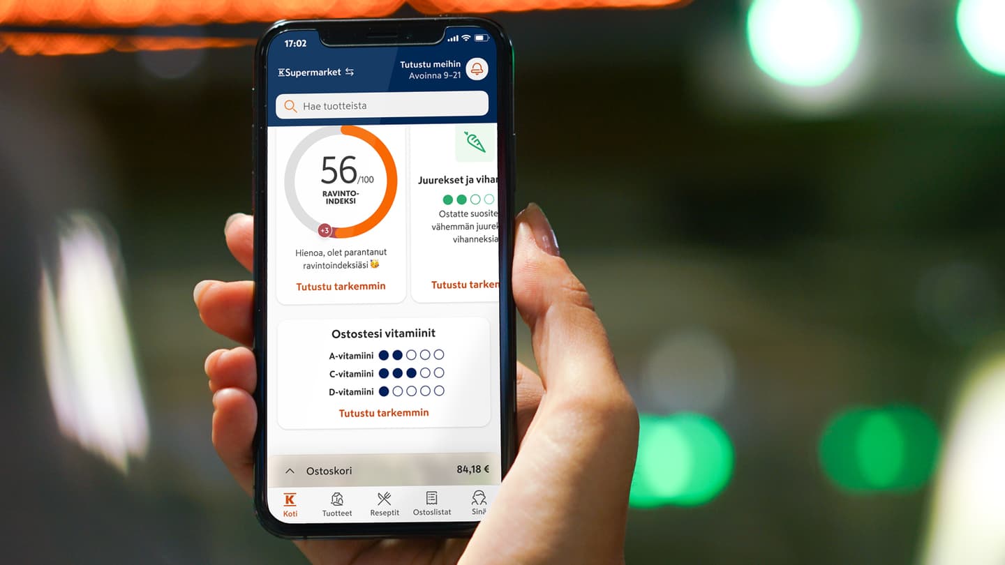 Hand holding smartphone displaying K-Supermarket app with nutrition index score of 56/100 and vitamin tracking features.