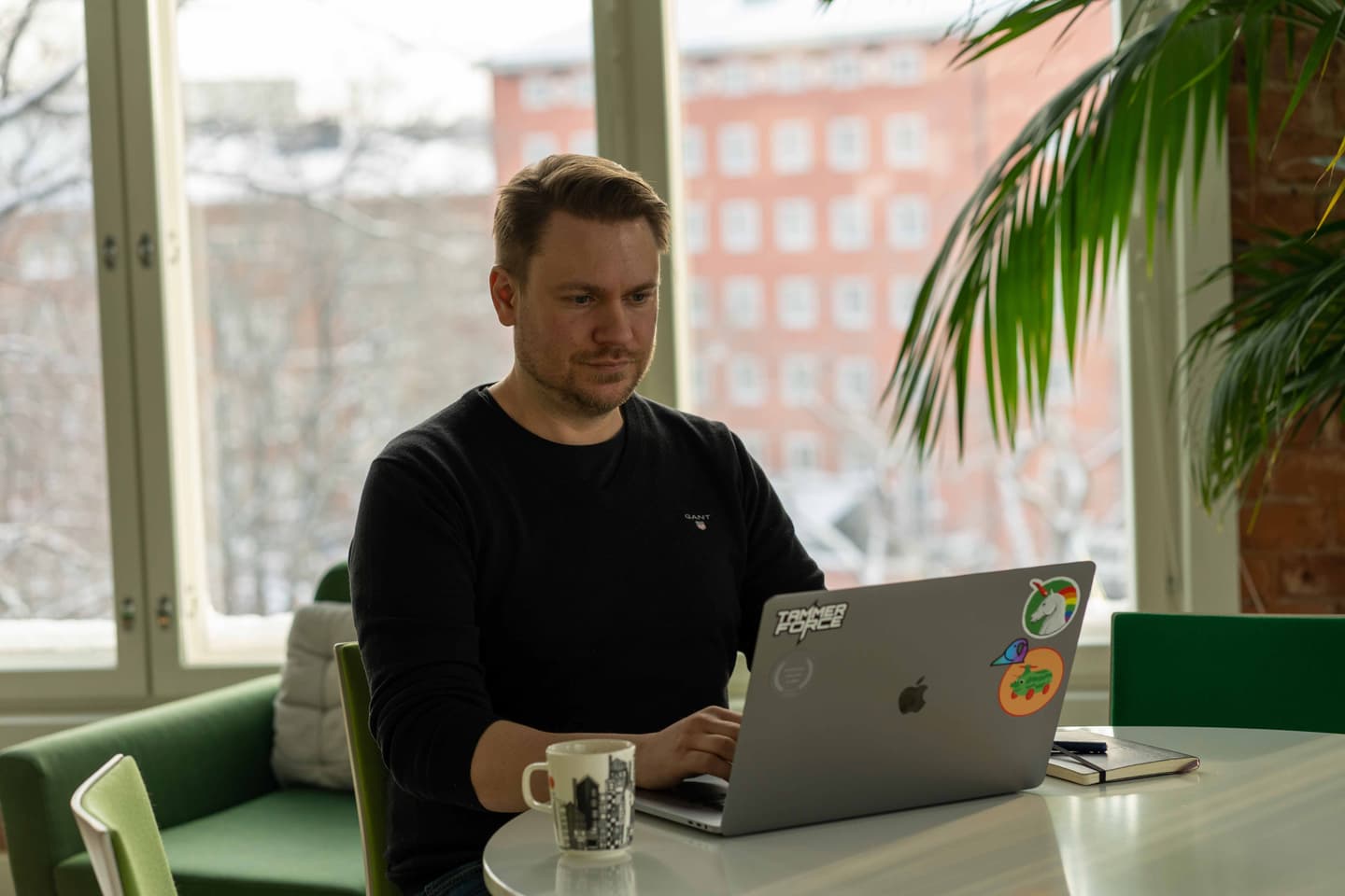 Person in black jumper working on laptop with stickered cover, coffee mug nearby, bright window view of brick building.