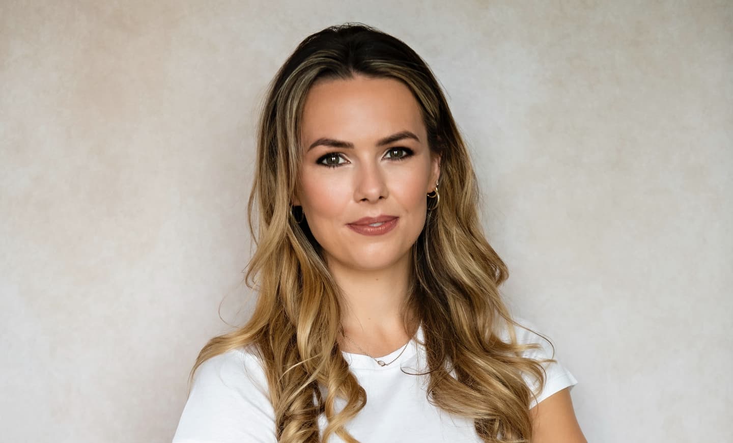 Portrait of a person with long wavy blonde hair wearing a white top against a neutral beige background.