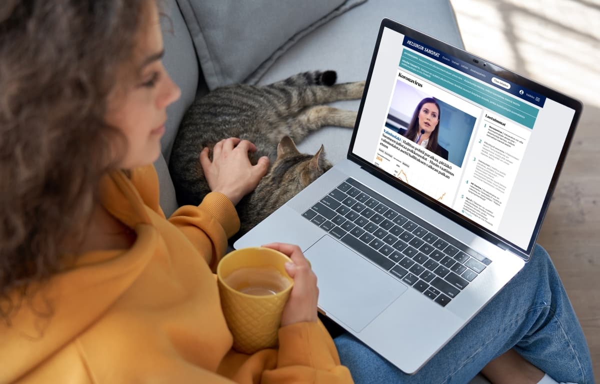 Person in orange jumper browsing Helsinki Sanomat website on laptop while petting cat and holding yellow mug.